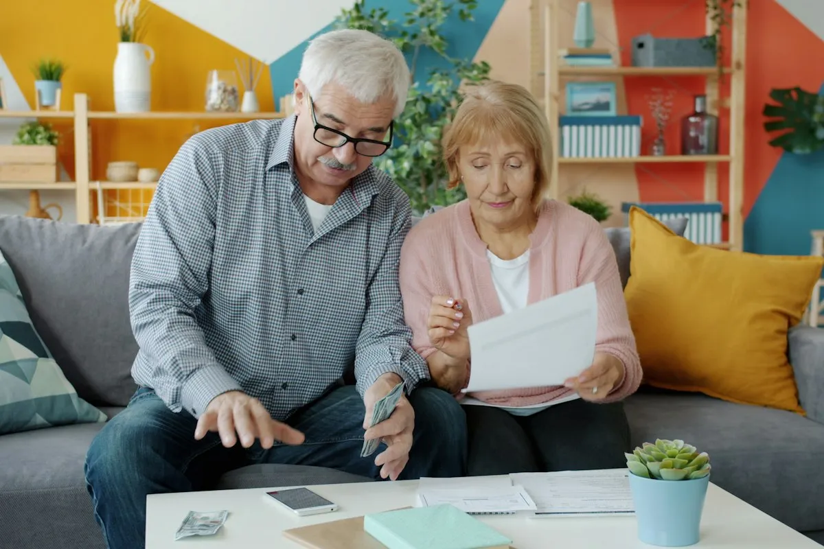 Older couple on a gray sofa reviewing paperwork together, the man in glasses holding a few banknotes and the woman in a pink cardigan reading a printed letter
