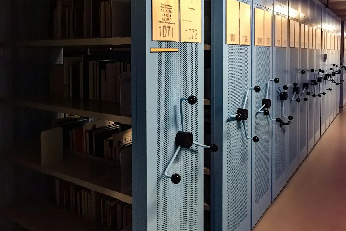 Rows of blue-gray mobile archive shelving in a dim records room, each bay fronted by a black crank handle and a numbered label card lit warm gold, with one bay rolled open to show shelves of upright bound volumes and box files
