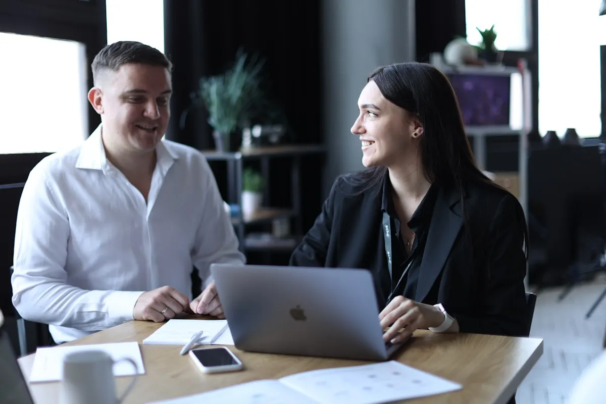 Two coworkers seated at a wooden desk in an office, smiling and talking to each other over an open laptop, a notebook, and a coffee mug
