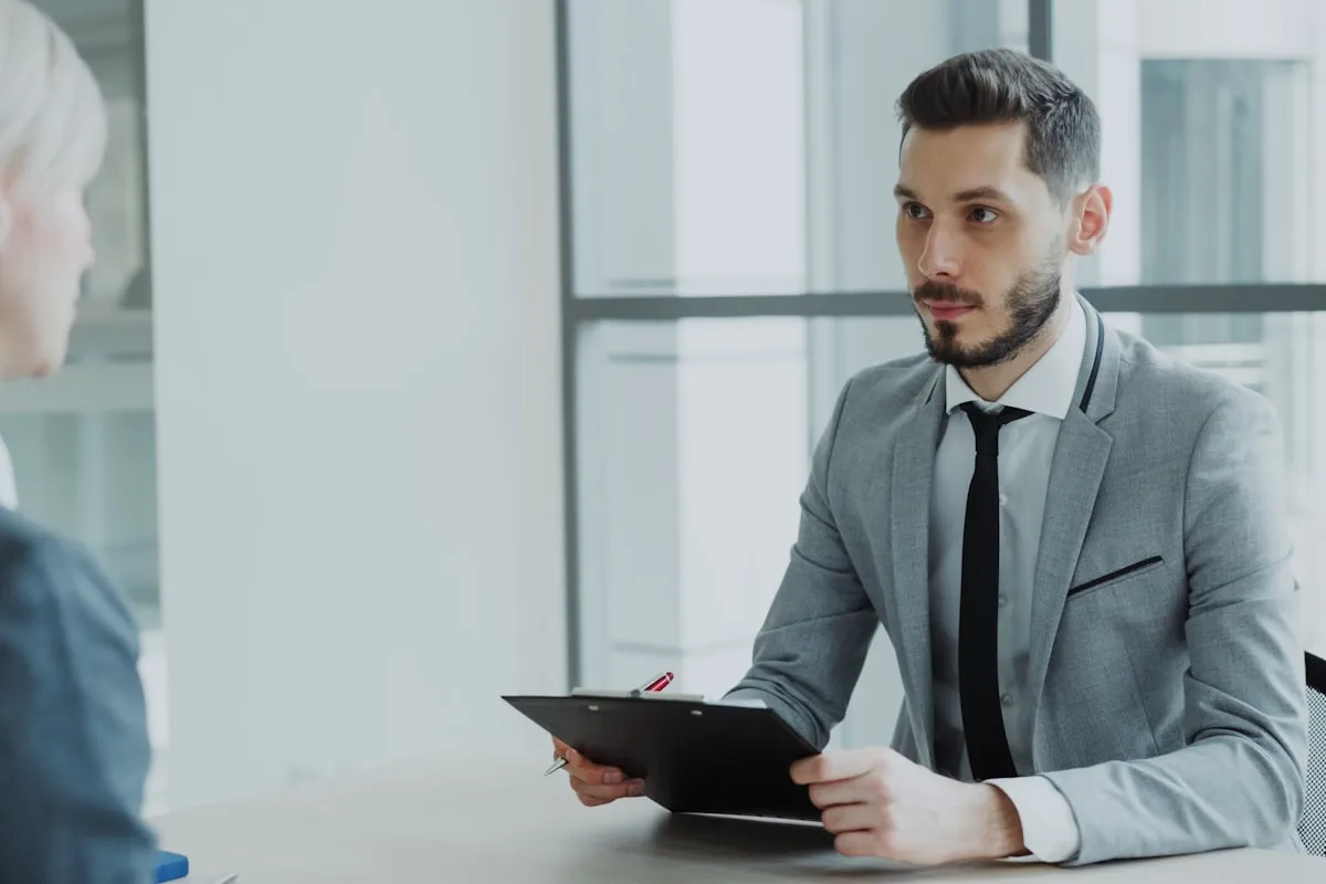 A bearded interviewer in a gray suit and dark tie holds a black clipboard and pen across a pale wood table from a candidate, in a bright office with glass partitions