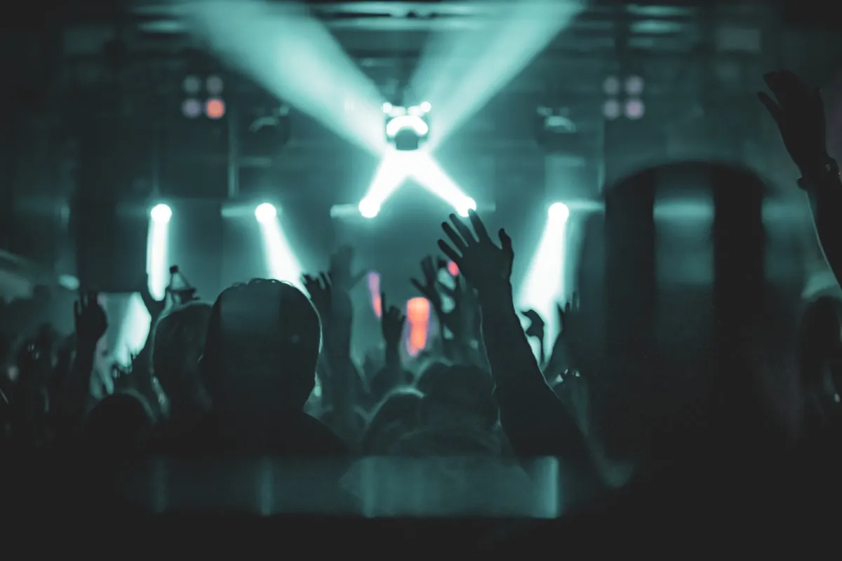 Concert crowd with hands raised in front of a brightly lit teal stage, fan silhouettes backlit by spotlights at a packed live show