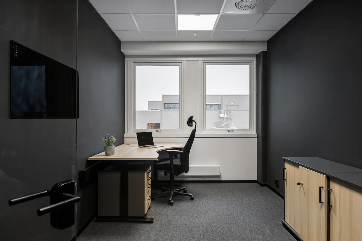 An unoccupied black office chair sits at a light wood desk holding an open laptop and a small potted plant, beside a bright window in a dark gray office.
