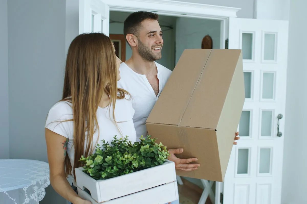 A smiling couple in white T-shirts carrying a cardboard moving box and a wooden crate of green plants through the open doorway of a new home