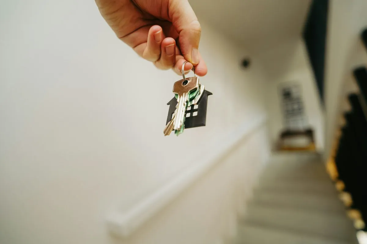 A hand dangling a set of keys on a black house-shaped keyring above a blurred, softly lit stairwell with a white handrail