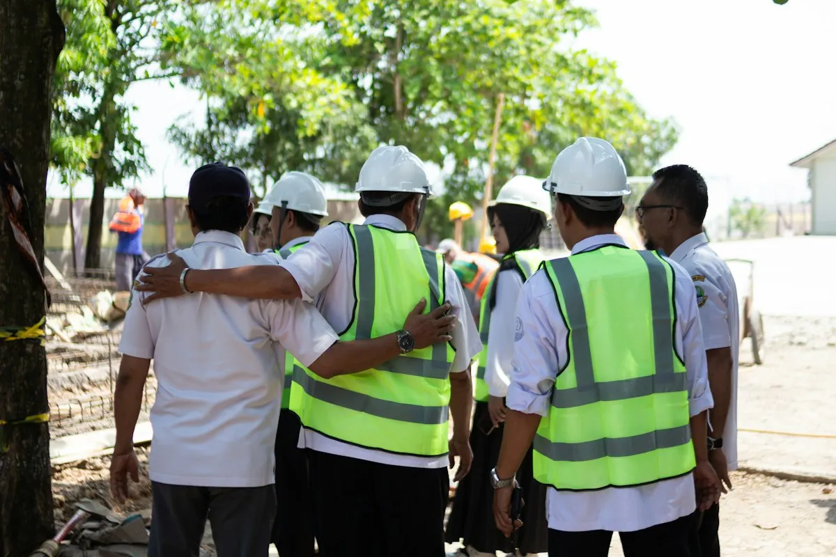 Workers, most in white hard hats and several in fluorescent yellow safety vests, standing together on a sunny construction site, one with an arm around a coworker's shoulders, seen from behind, with rebar mesh and trees in the background