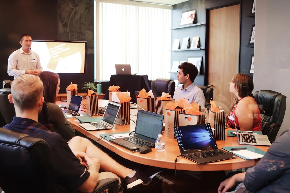 A presenter stands beside a screen leading a training session for a team seated around a conference table with open laptops in an office.