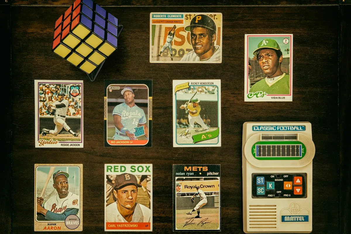 Eight vintage baseball cards arranged in three rows on dark stained wood, with a colored puzzle cube in the top corner and a Mattel handheld electronic football game at the right