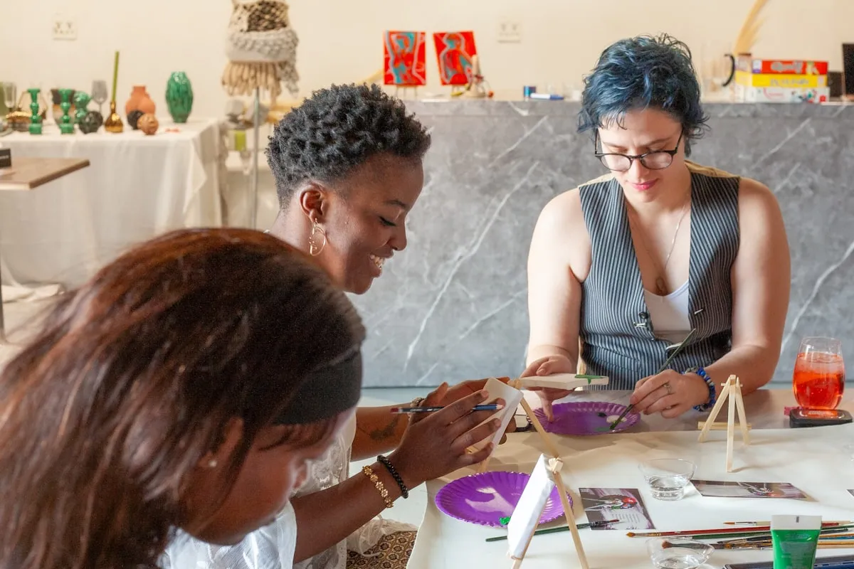 Three women in summer clothing painting small canvases together at a studio worktable, with handmade pottery and craft supplies on shelves behind them