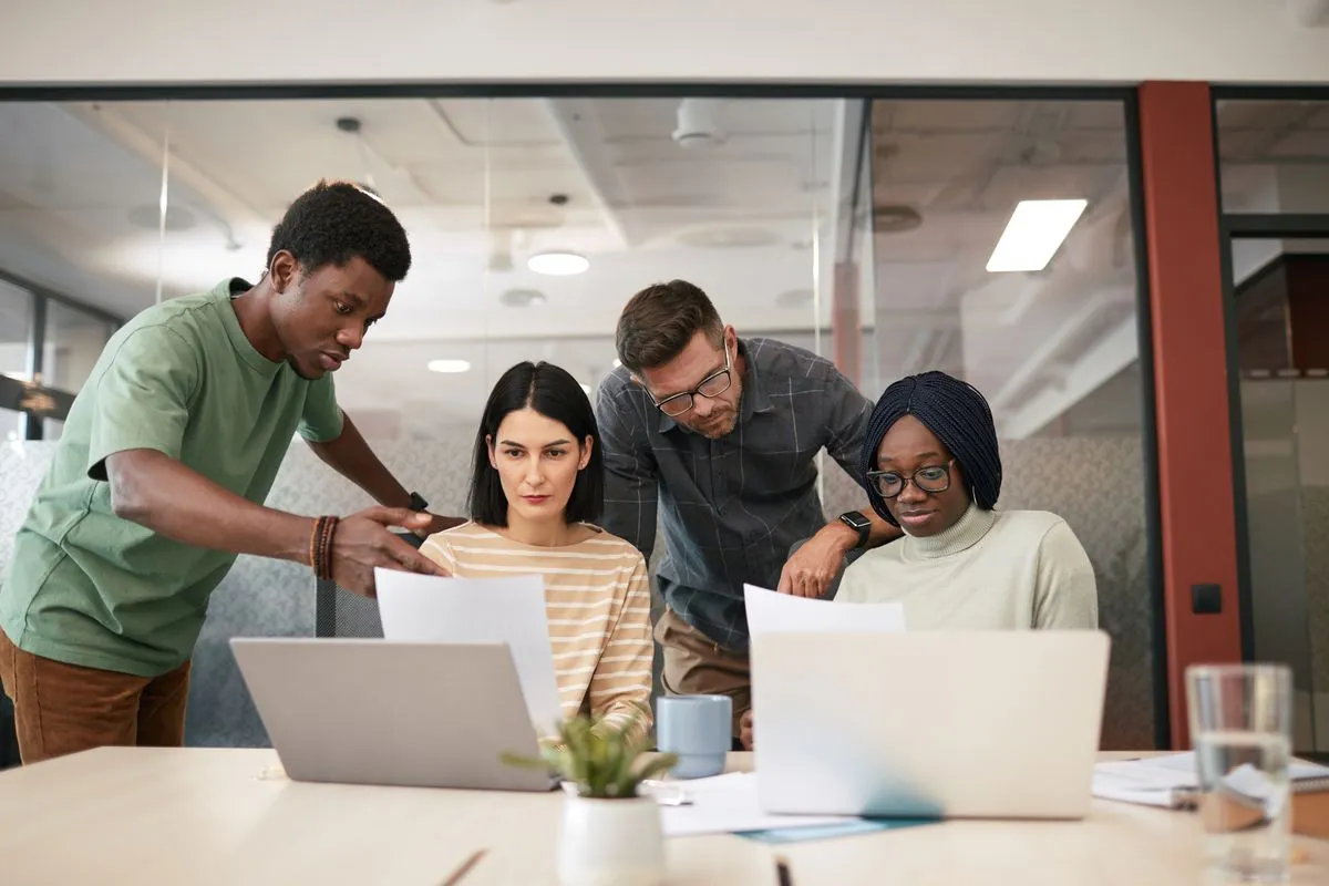 Four coworkers gathered around a meeting table with two open laptops, one man pointing at a printed page while another sheet is held up across the table, in a glass-walled office.