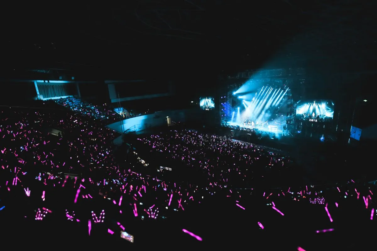 A darkened concert arena seen from the upper seats, the crowd holding hundreds of glowing pink penlights toward a stage lit by blue spotlights and two video screens