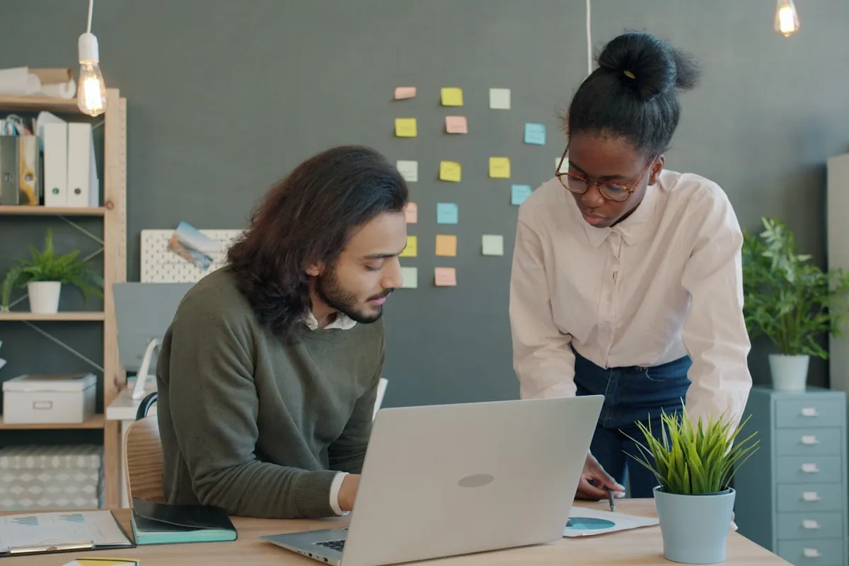 Two coworkers at a desk reviewing work on a laptop together, one seated and typing while the other stands pointing at a printed chart, with sticky notes on the wall behind them