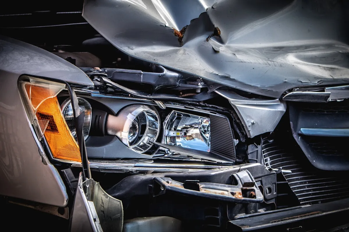 Close-up of the crushed front end of a silver car, showing a crumpled hood, a shattered headlight assembly with exposed bulbs, and a torn-away bumper