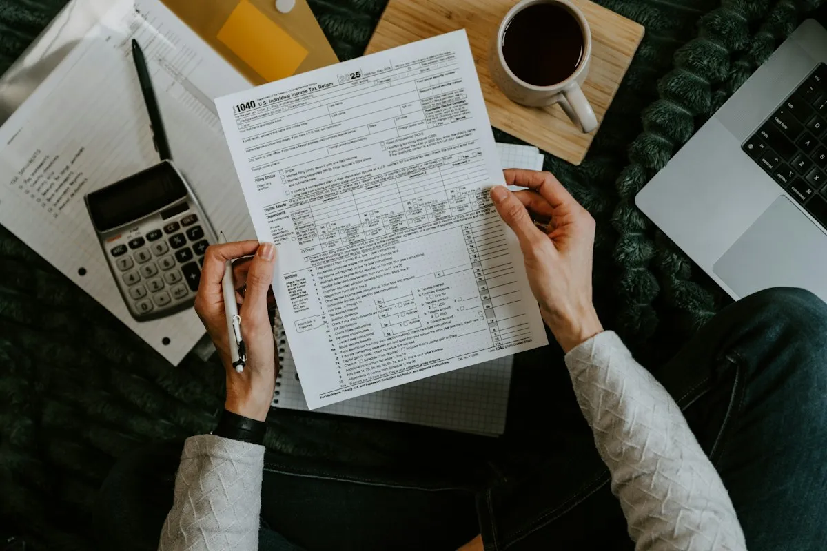 Overhead view of a person in a cream sweater holding a pen and a printed Form 1040 tax return, with a calculator, notebook, laptop and mug of coffee on a dark green blanket