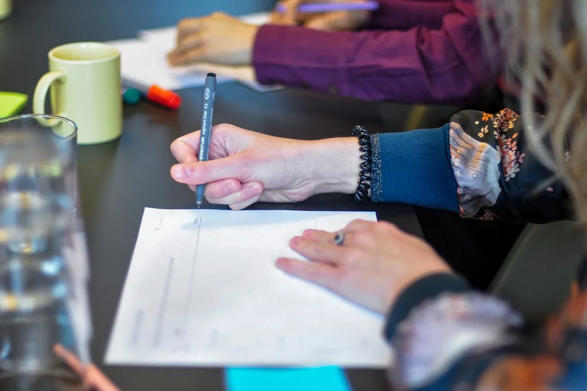 Two pairs of hands at a dark meeting table, one holding a fineliner over a printed form, with a pale green mug and a water jug beside them