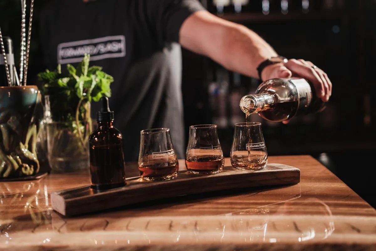 A bartender pours an amber spirit into the third of three tulip-shaped tasting glasses lined up on a wooden flight board, with a dropper bottle and fresh mint on a polished bar top